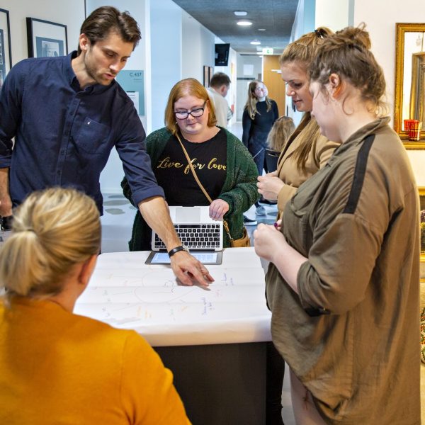 Fire studerende og en lærer er samlet om et bord, og læreren er i færd med at forklare noget.