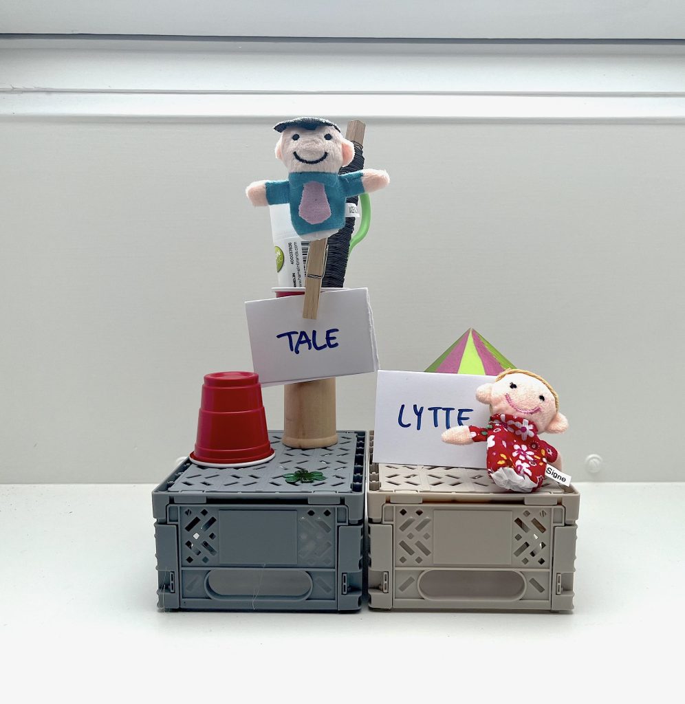 En installation med forskellige figurer på kasser. Til hver figur er der et stykke papir. På det ene står der "tale", og på det andet står der "lytte".