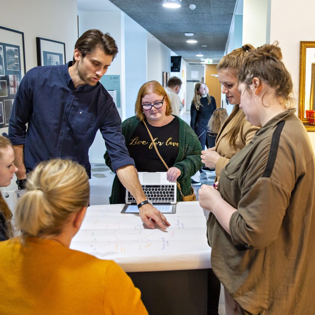 Fire studerende og en lærer er samlet om et bord, og læreren er i færd med at forklare noget.